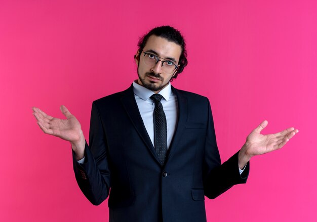 Hombre de negocios en traje negro y gafas mirando al frente con las manos levantadas con expresión confusa de pie sobre la pared rosa