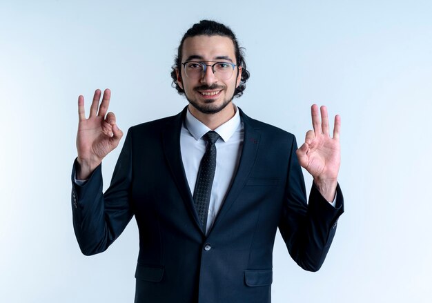 Hombre de negocios en traje negro y gafas mirando al frente mostrando el signo de ok con ambas manos sonriendo de pie sobre la pared blanca