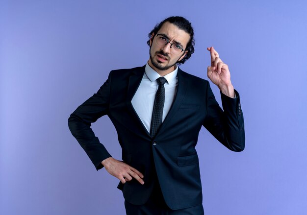 Hombre de negocios en traje negro y gafas mirando al frente pidiendo deseo cruzando los dedos de pie sobre la pared azul