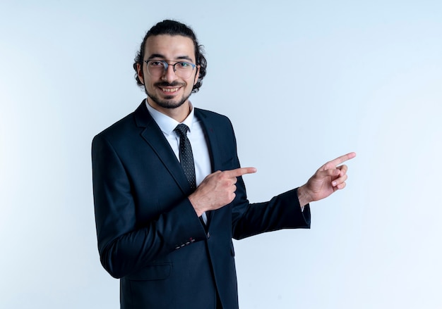 Hombre de negocios en traje negro y gafas mirando al frente sonriendo alegremente apuntando con los dedos índices hacia el lado de pie sobre la pared blanca