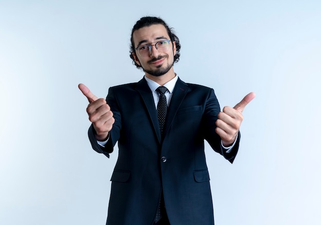 Hombre de negocios en traje negro y gafas mirando al frente sonriendo alegremente mostrando los pulgares para arriba de pie sobre la pared blanca