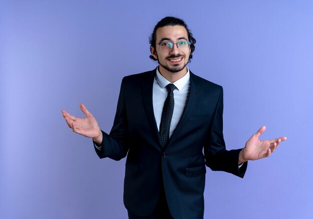 Hombre de negocios en traje negro y gafas mirando al frente sonriendo amable haciendo gesto de bienvenida con las manos de pie sobre la pared azul