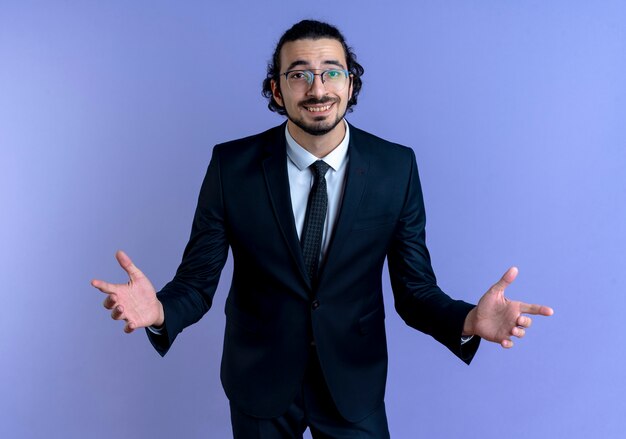 Hombre de negocios en traje negro y gafas mirando al frente sonriendo haciendo gesto de bienvenida con las manos de pie sobre la pared azul