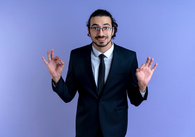 Hombre de negocios en traje negro y gafas mirando al frente sonriendo haciendo gesto de meditación con los dedos de pie sobre la pared azul