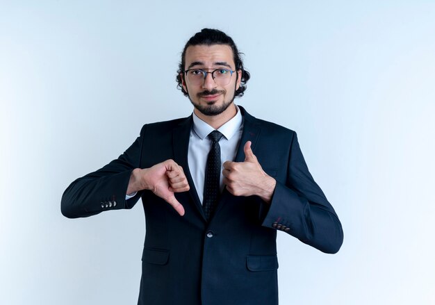 Hombre de negocios en traje negro y gafas mirando al frente con una sonrisa escéptica mostrando los pulgares hacia arriba y hacia abajo de pie sobre la pared blanca