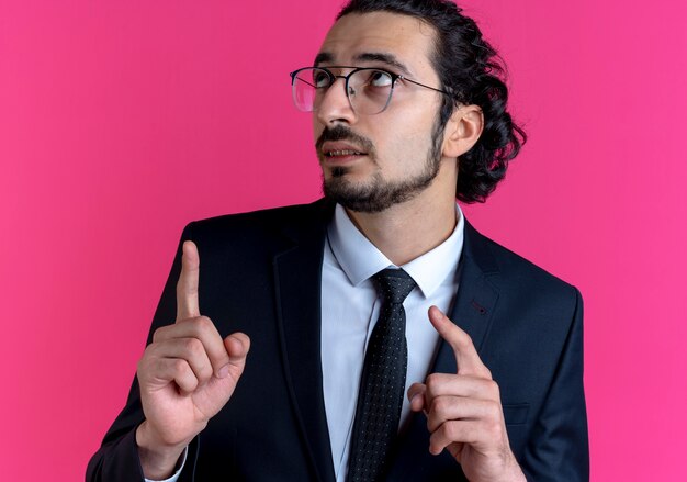 Hombre de negocios en traje negro y gafas mirando hacia arriba apuntando con los dedos hacia el lado parado sobre la pared rosa