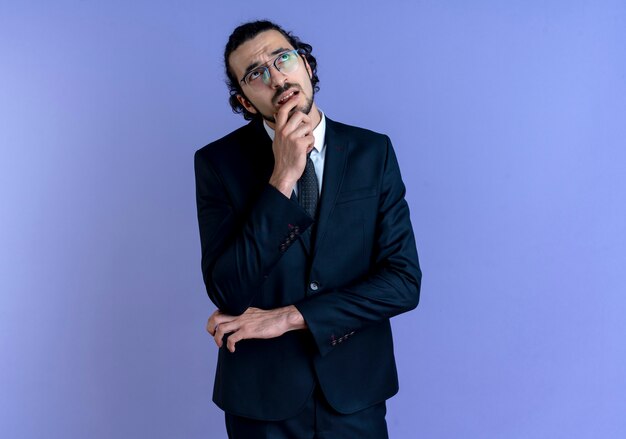 Hombre de negocios en traje negro y gafas mirando hacia arriba con la mano en la barbilla, desconcertado de pie sobre la pared azul