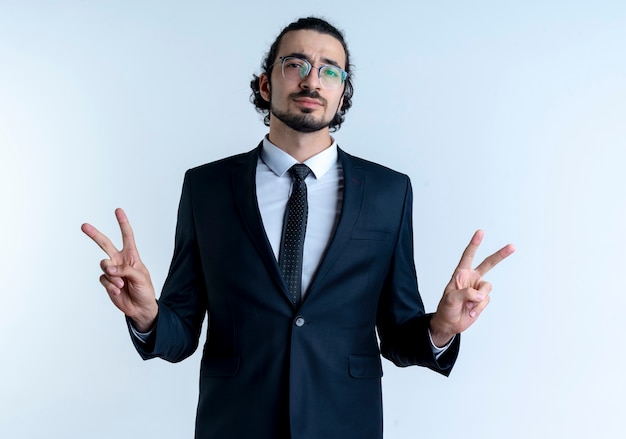 Hombre de negocios en traje negro y gafas mirando confiado mostrando el signo de la victoria con ambas manos de pie sobre la pared blanca