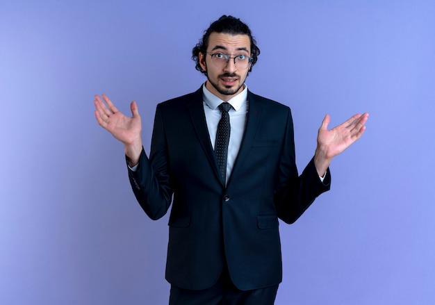 Hombre de negocios en traje negro y gafas mirando confundido encogiéndose de hombros sin respuesta de pie sobre la pared azul