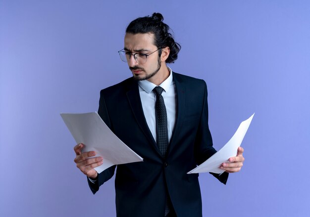 Hombre de negocios en traje negro y gafas mirando documentos con cara seria de pie sobre la pared azul