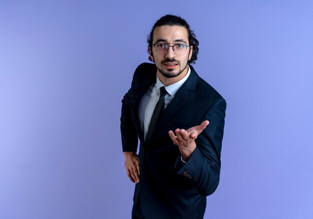 Hombre de negocios en traje negro y gafas mirando hacia el frente con el brazo extendido como haciendo una pregunta de pie sobre la pared azul