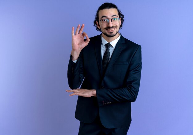 Hombre de negocios en traje negro y gafas mirando hacia el frente sonriendo alegremente mostrando signo ok de pie sobre la pared azul