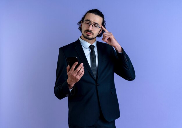 Hombre de negocios en traje negro y gafas mirando la pantalla de su teléfono inteligente apuntando con el dedo a su sien se centró en una tarea de pie sobre la pared azul