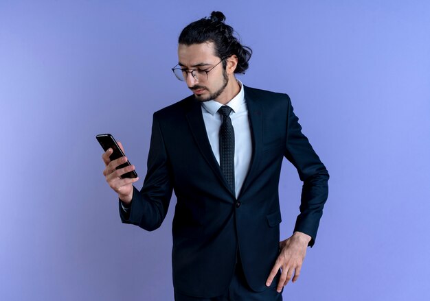 Hombre de negocios en traje negro y gafas mirando la pantalla de su teléfono inteligente con cara seria de pie sobre la pared azul