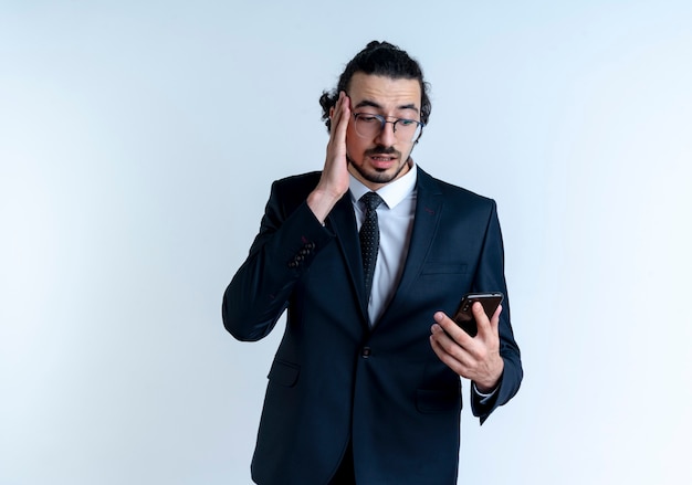 Hombre de negocios en traje negro y gafas mirando la pantalla de su teléfono inteligente confundido y muy ansioso de pie sobre la pared blanca
