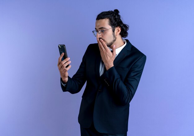 Hombre de negocios en traje negro y gafas mirando la pantalla de su teléfono inteligente sorprendido y confundido de pie sobre la pared azul