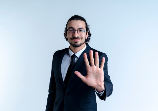 Hombre de negocios en traje negro y gafas mostrando y apuntando hacia arriba con los dedos número cinco sonriendo de pie sobre la pared blanca