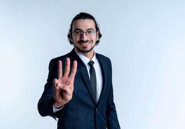 Hombre de negocios en traje negro y gafas mostrando y apuntando hacia arriba con los dedos número tres sonriendo de pie sobre la pared blanca