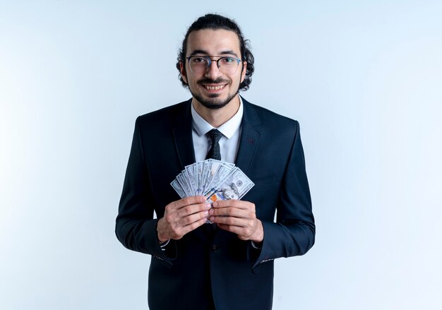 Hombre de negocios en traje negro y gafas mostrando dinero en efectivo mirando al frente sonriendo alegremente de pie sobre la pared blanca