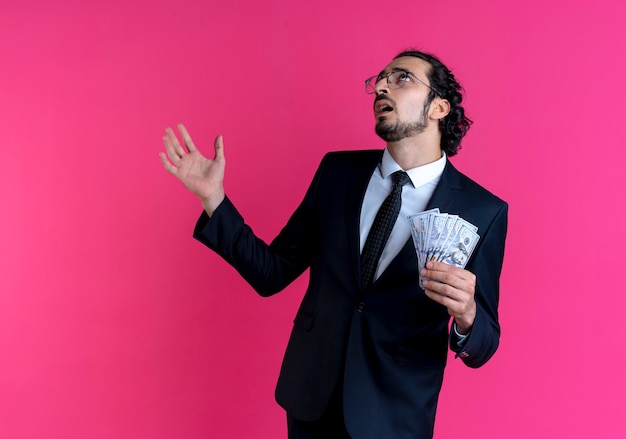 Hombre de negocios en traje negro y gafas mostrando dinero en efectivo, mirando hacia arriba confundido con el brazo de pie sobre la pared rosa