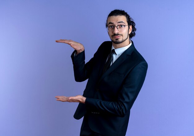 Hombre de negocios en traje negro y gafas mostrando gesto de tamaño con las manos, símbolo de medida de pie sobre la pared azul