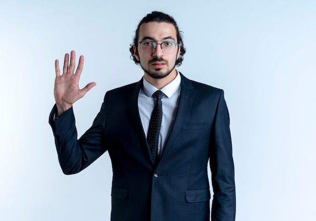 Hombre de negocios en traje negro y gafas mostrando la mano abierta con cara seria de pie sobre la pared blanca