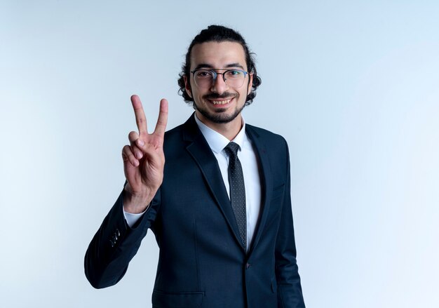 Hombre de negocios en traje negro y gafas mostrando el signo de la victoria sonriendo alegremente de pie sobre la pared blanca