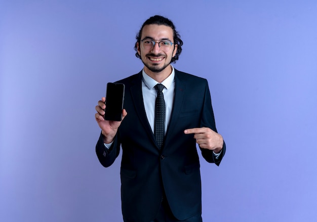 Hombre de negocios en traje negro y gafas mostrando smartphone apuntando con el dedo sonriendo confiados de pie sobre la pared azul