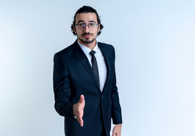 Hombre de negocios en traje negro y gafas ofreciendo saludo de mano mirando confiado de pie sobre la pared blanca