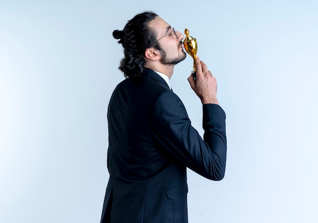 Hombre de negocios en traje negro y gafas de pie lateralmente besando su trofeo sobre la pared blanca