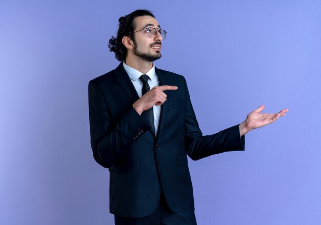 Hombre de negocios en traje negro y gafas presentando con el brazo de su mano apuntando con el dedo hacia el lado mirando confiado de pie sobre la pared azul