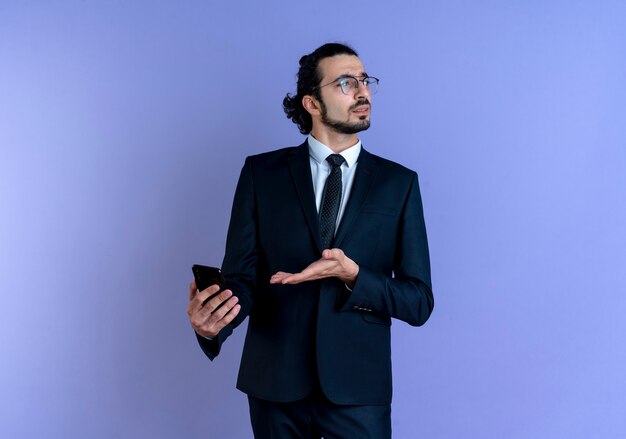 Hombre de negocios en traje negro y gafas con smartphone mirando confundido parado sobre pared azul
