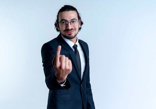 Hombre de negocios en traje negro y gafas sonriendo alegremente mostrando el dedo índice de pie sobre la pared blanca