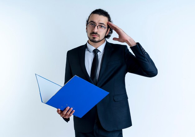 Hombre de negocios en traje negro y gafas sosteniendo una carpeta mirando hacia el frente con expresión de confianza saludando de pie sobre la pared blanca