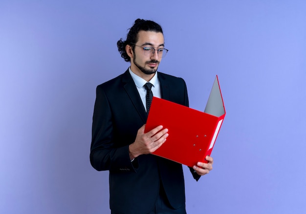 Hombre de negocios en traje negro y gafas sosteniendo una carpeta roja mirándolo con cara seria de pie sobre la pared azul