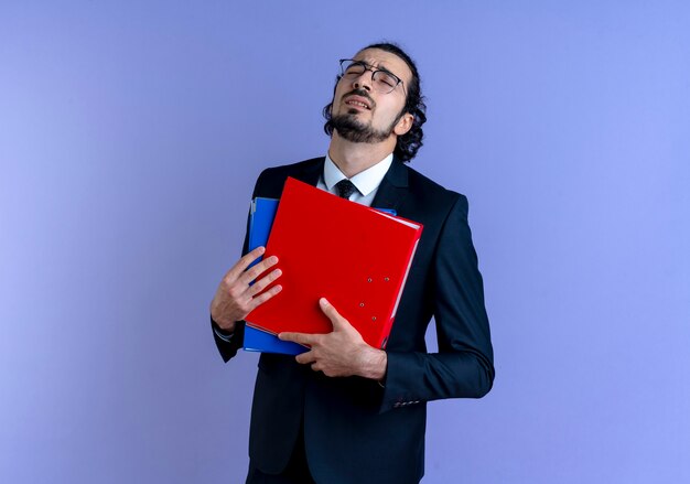 Hombre de negocios en traje negro y gafas sosteniendo carpetas con los ojos cerrados cansado y aburrido de pie sobre la pared azul