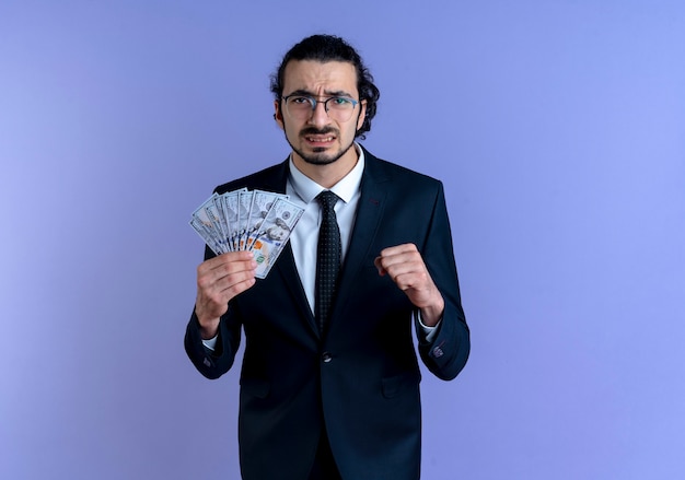 Hombre de negocios en traje negro y gafas sosteniendo dinero en efectivo mirando al frente apretando el puño con expresión molesta de pie sobre la pared azul