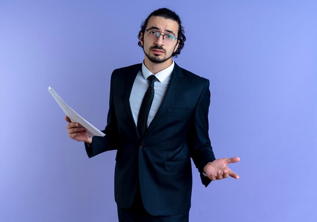 Hombre de negocios en traje negro y gafas sosteniendo documentos mirando hacia el frente confundido de pie sobre la pared azul