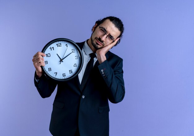 Hombre de negocios en traje negro y gafas sosteniendo un reloj de pared con aspecto cansado y con exceso de trabajo haciendo gesto de sueño de pie sobre la pared azul