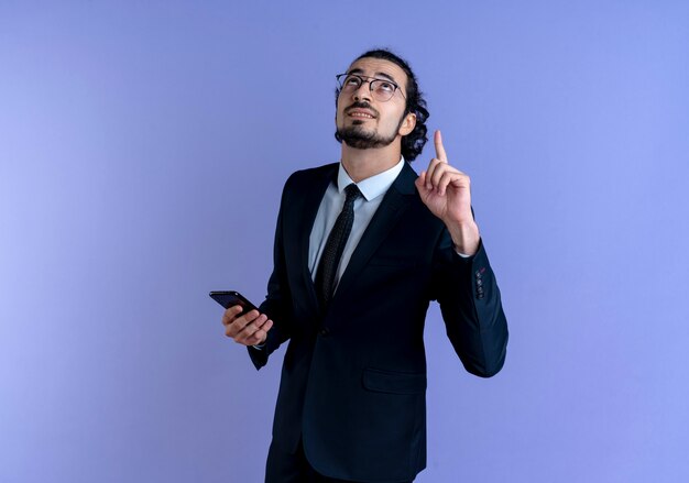 Hombre de negocios en traje negro y gafas sosteniendo smartphone apuntando con el dedo índice hacia arriba mirando confiado de pie sobre la pared azul