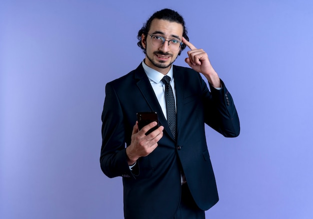 Hombre de negocios en traje negro y gafas sosteniendo smartphone apuntando con el dedo su sien mirando confundido parado sobre pared azul