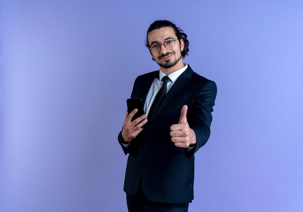 Hombre de negocios en traje negro y gafas sosteniendo smartphone mostrando Thumbs up sonriendo confiados de pie sobre la pared azul