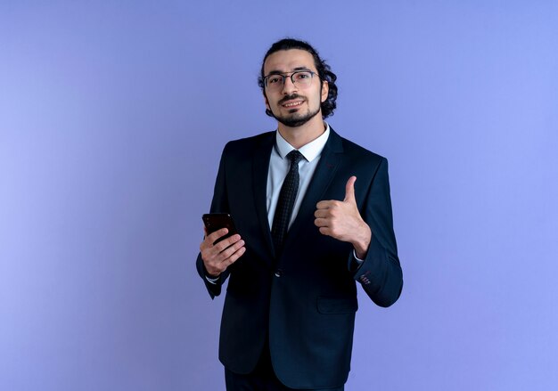 Hombre de negocios en traje negro y gafas sosteniendo smartphone mostrando Thumbs up sonriendo de pie sobre la pared azul