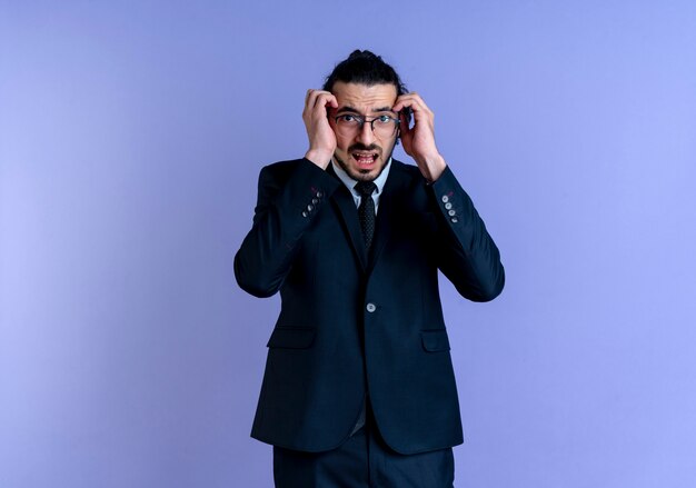 Hombre de negocios en traje negro y gafas sosteniendo su cabeza con las manos mirando hacia el frente con expresión de miedo de pie sobre la pared azul