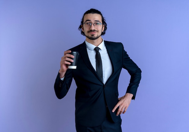 Hombre de negocios en traje negro y gafas sosteniendo la taza de café mirando hacia el frente con expresión de confianza de pie sobre la pared azul