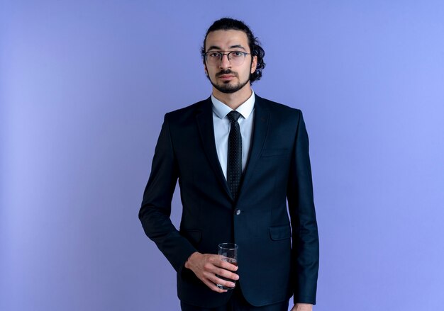 Hombre de negocios en traje negro y gafas sosteniendo un vaso de agua mirando hacia el frente con cara seria de pie sobre la pared azul