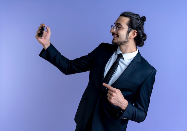 Hombre de negocios en traje negro y gafas con tarjeta de crédito apuntando con el dedo hacia el lado sonriendo alegremente de pie sobre la pared azul