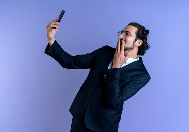 Hombre de negocios en traje negro y gafas tomando selfie con su teléfono inteligente mirando al frente con una sonrisa tímida en la cara de pie sobre la pared azul