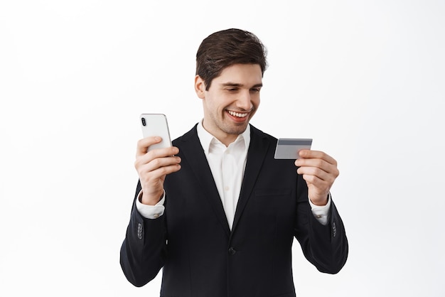 Foto gratuita hombre de negocios con traje negro mira la tarjeta de crédito mientras paga en línea con compras de teléfonos móviles en línea en la aplicación de teléfono inteligente pide algo de fondo blanco sin contacto