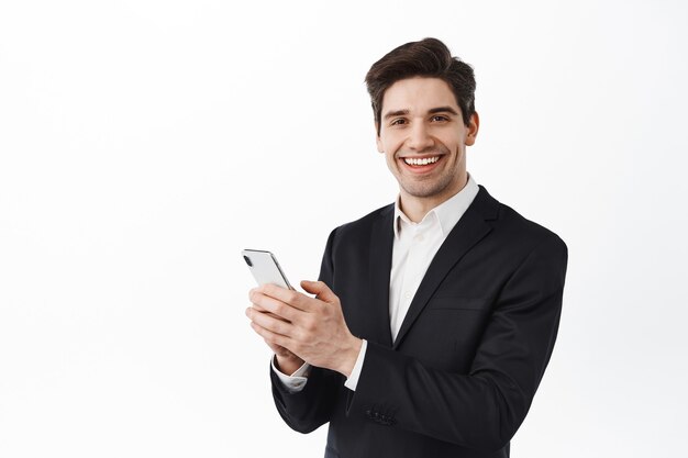 Hombre de negocios en traje negro mediante teléfono móvil, de pie con smartphone y mirando al frente, sonriendo, pared blanca